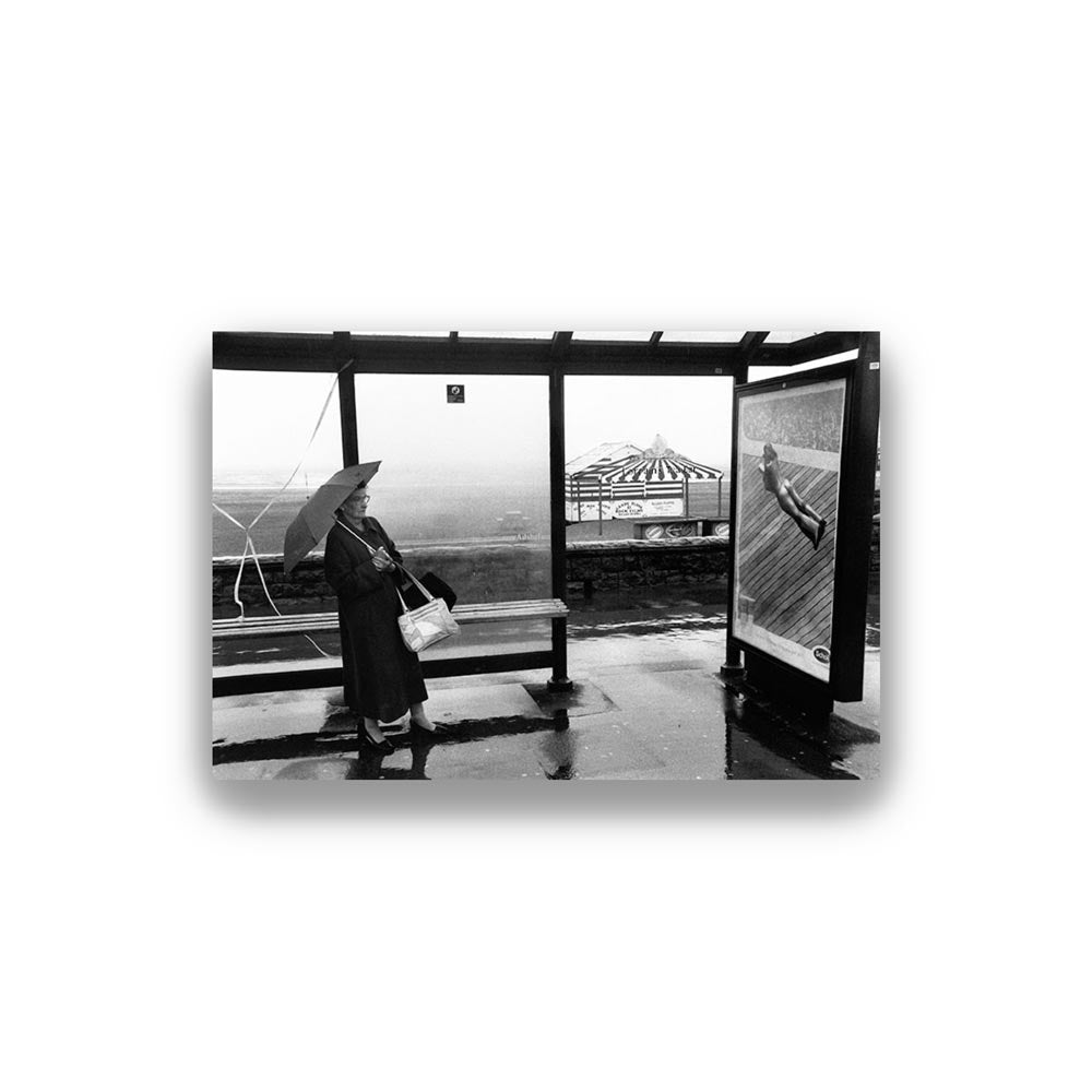 Black and white photo of a person with an umbrella at a bus stop. Photo by Sir Roger Deakins