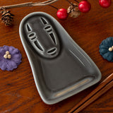 Gray ceramic tray on a wooden surface with decorative elements. Ceramic condiment dish in the shape of No Face from Studio Ghibli's Spirited Away.