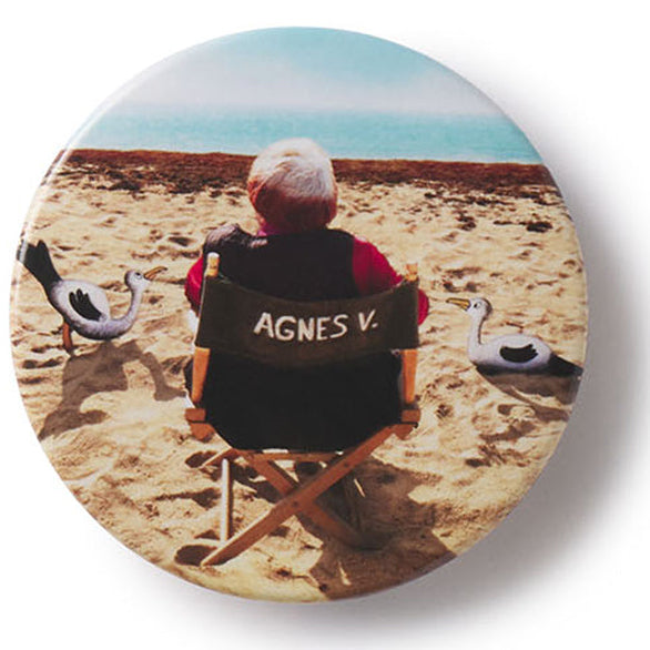 Button badge with film director Agnes Varda on a beach, wearing a jacket with 'AGNES V.' text.