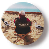 Button badge with film director Agnes Varda on a beach, wearing a jacket with 'AGNES V.' text.