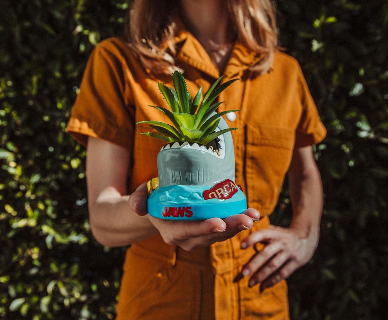 Person holding a unique plant pot shaped like a wave with 'Jaws' branding against a green leafy background.