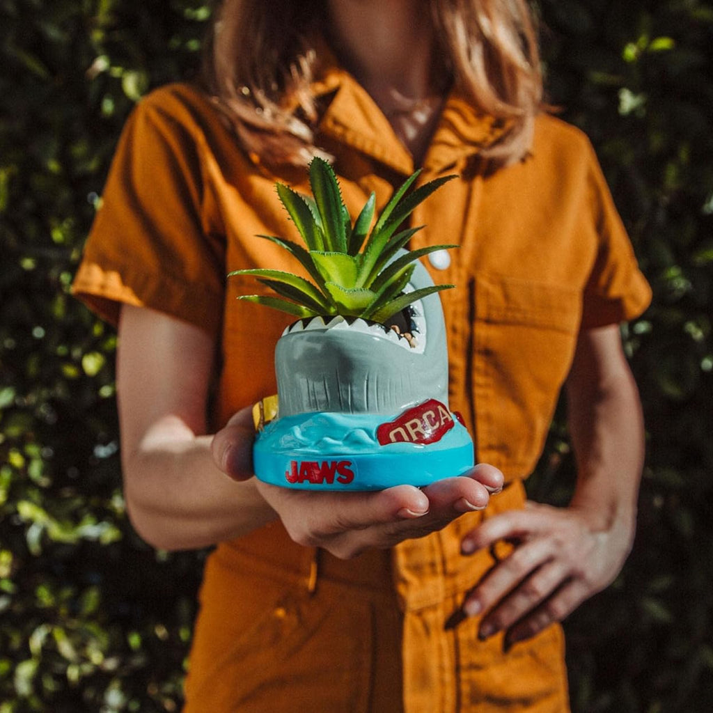 Person holding a unique plant pot shaped like a wave with 'Jaws' branding against a green leafy background.