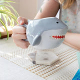Person holding a shark-shaped mug with a tea bag inside on a table.