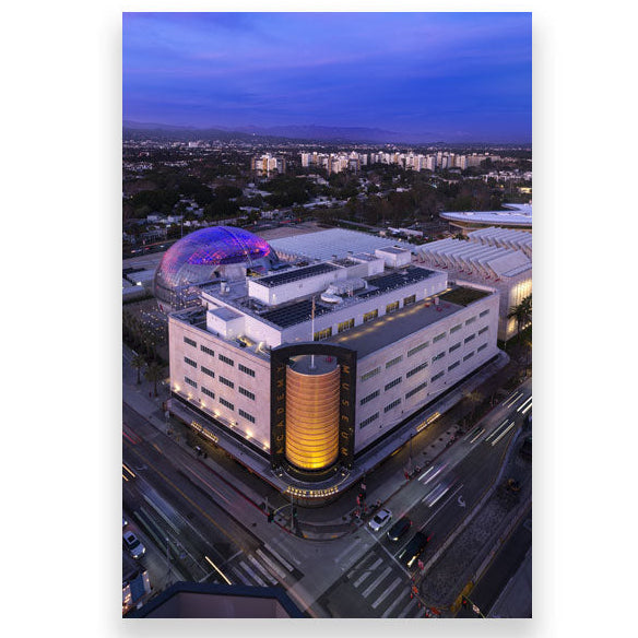 Modern building with a unique design at dusk, surrounded by a cityscape. Academy Museum Postcard Aerial View The Gold Cylinder Facade Size 4 x 6in. Academy Museum Store Exclusive  
©Academy Museum Foundation, Photo by: Fredrik Nilsen
