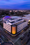 Aerial view of a modern building with a distinctive design at dusk. Academy Museum Postcard Aerial View The Gold Cylinder Facade Size 4 x 6in. Academy Museum Store Exclusive  
©Academy Museum Foundation, Photo by: Fredrik Nilsen
