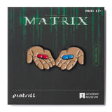Enamel pin of hands holding red and blue pills from 'The Matrix' on a black background with 'Pintrill' and 'Academy Museum' logos.