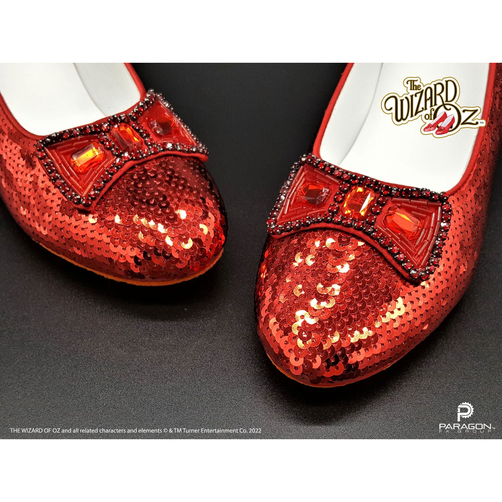 Red glittery shoes with decorative elements on a black background, featuring 'The Wizard of Oz' branding.Prop Replica Authentic Dorothy's Ruby Slippers from The Wizard of Oz