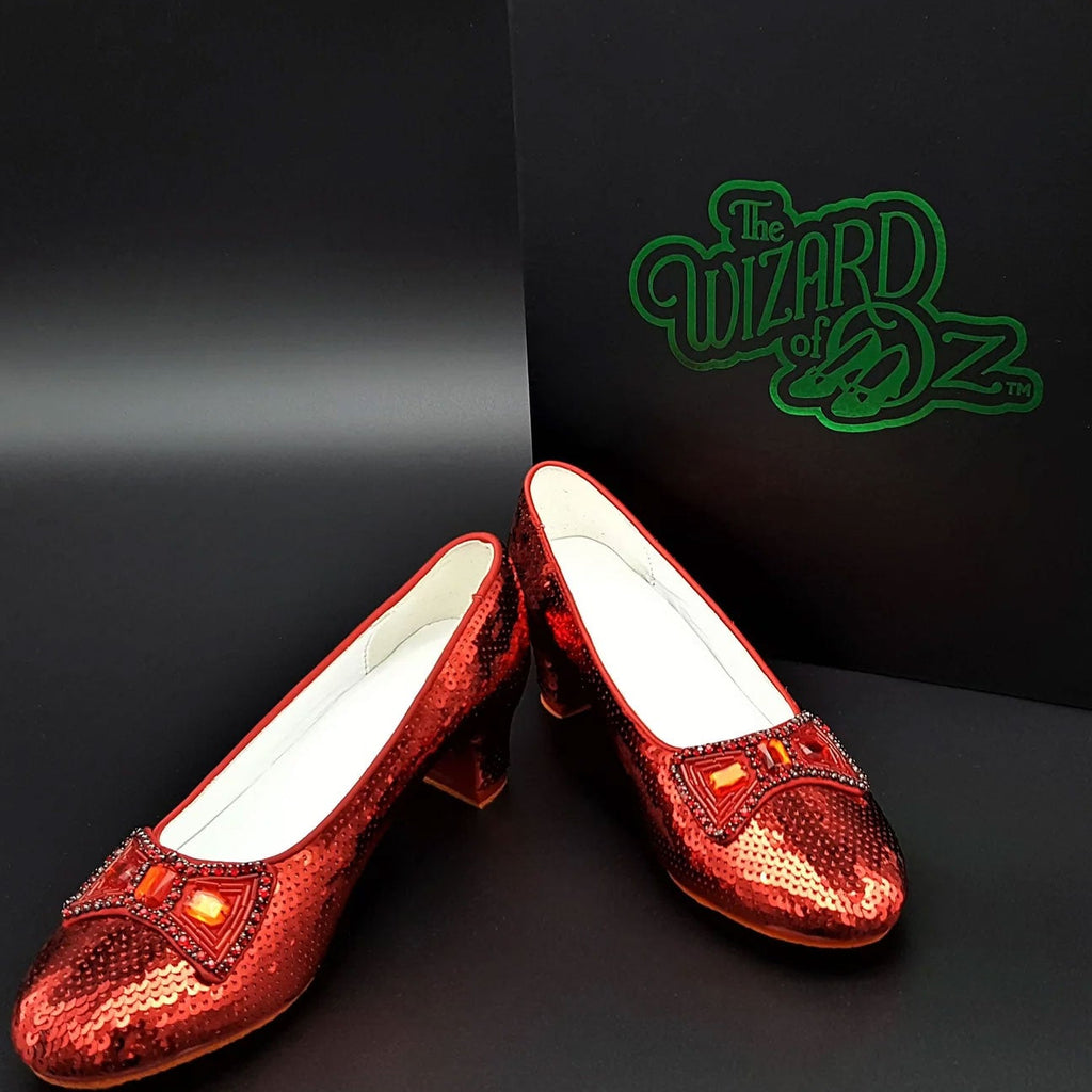 Red glittery shoes with white soles in front of a 'The Wizard of Oz' branded box on a dark background. Movie Prop Authentic Museum Quality Replica Dorothy's Ruby Slippers from The Wizard of Oz
