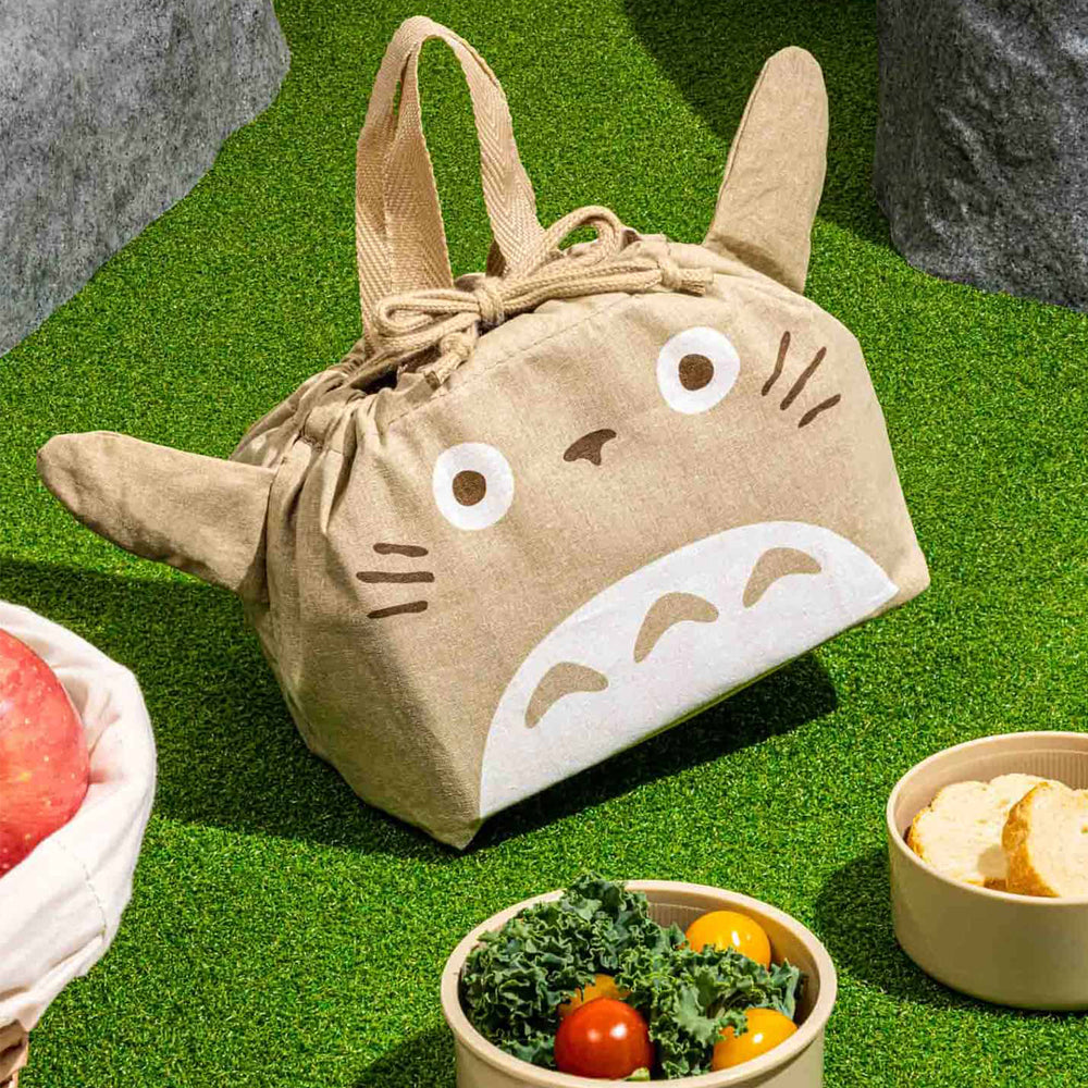 Tan drawstring lunch bag with cartoon face design on grass with bowls of food