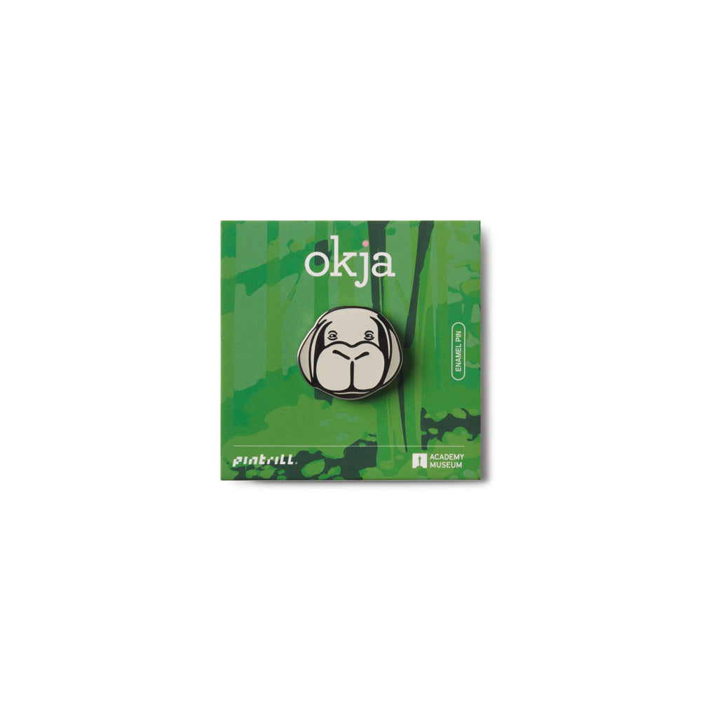 This is an enamel lapel pin on Green card with a logo and 'okja' text on a white background featuring the face of Okja from Bong Joon Ho's film Okja.