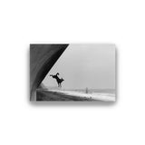 Black and white photo of a person standing on one leg by a body of water with a large archway.