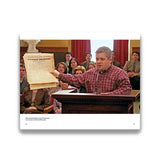 Man in a courtroom holding a document, with a group of people seated behind him.
