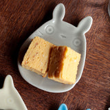 Two pieces of yellow cake on a white ceramic plate shaped like a Totoro character, on a wooden surface.