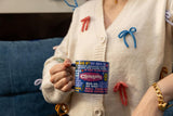 Person holding a Clueless mug with a colorful design, wearing a white sweater with colorful bows. Celebrate the 90s with our Clueless Coffee Mug! Officially licensed, it showcases memorable quotes and comes in a fun gift box. Perfect for any cinephile!