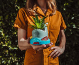 Person holding a unique plant pot shaped like a wave with 'Jaws' branding against a green leafy background.