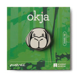 Enamel pin with 'okja' design on a green card with 'Pintrill' and 'Academy Museum' branding.