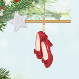 Red glittery shoes ornament hanging on a Christmas tree with a star and gold bar in the background. Bring home the magic of Oz with the Ruby Slippers™ ornament! This delightful keepsake is perfect for any fan, adding sparkle to your holiday celebrations.