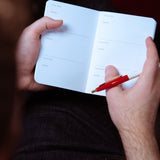 Person holding a small notebook and red pen with a dark background. Experience the art of film note-taking with our elegant Pocket Size Movie Screening Note Journal, inspired by Academy designs. Perfect for the discerning cinephile.
