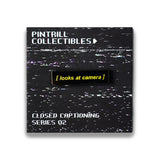 Pin with 'looks at camera' text on a black background with white specks, branded by Pintrill Collectibles.